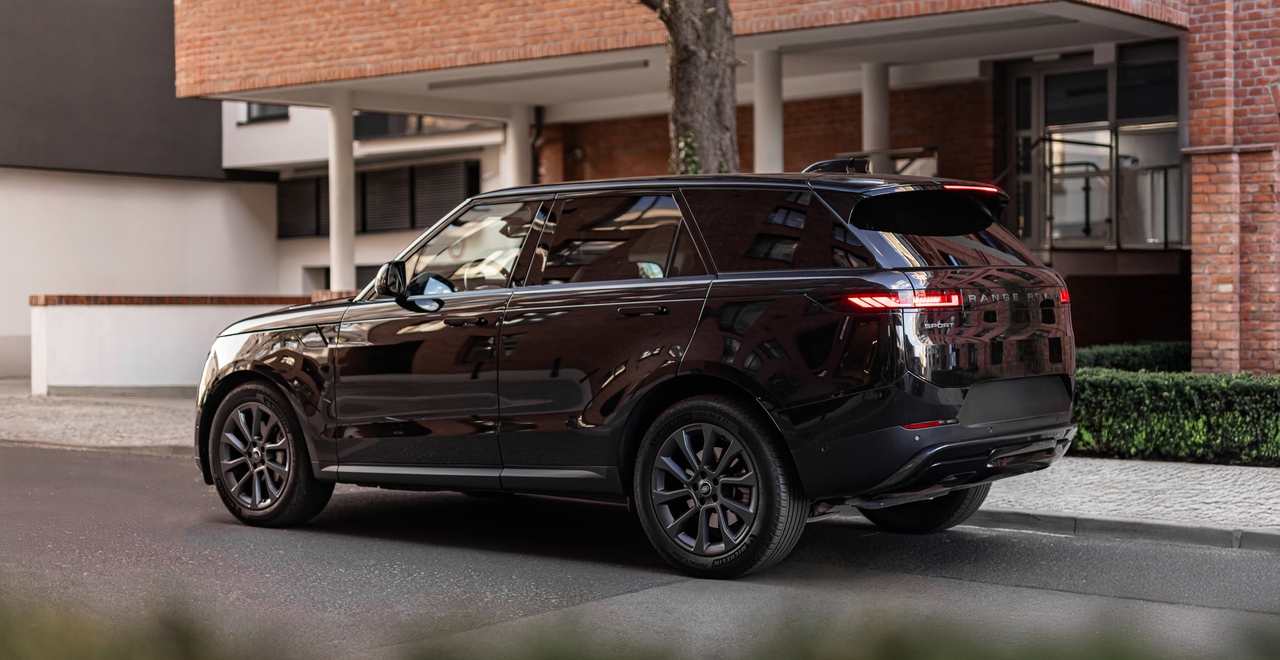 Czarny Range Rover Sport, widok tylno-boczny, zaparkowany na tle ceglanego budynku i nowoczesnej elewacji. Na karoserii wyraźnie odbijają się okoliczne budynki.