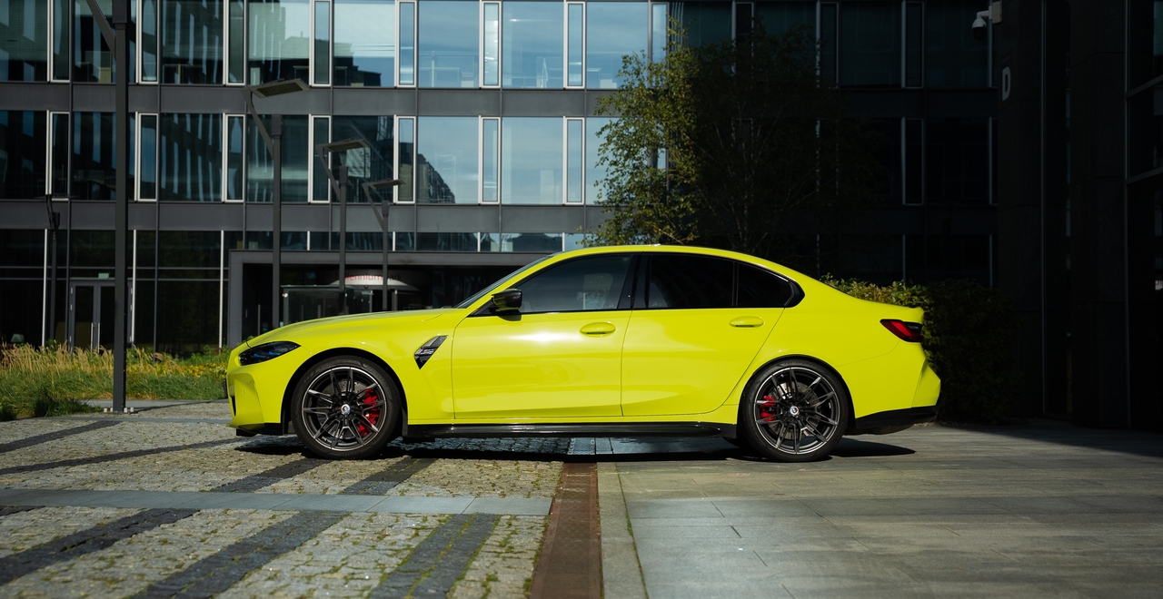 Jaskrawożółte BMW M3 Competition xDrive z profilu, na tle nowoczesnego biurowca.
