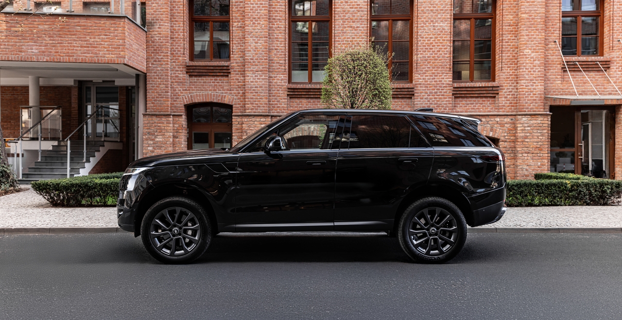 Czarny Range Rover Sport z profilu, zaparkowany na tle ceglanego budynku z dużymi oknami i krzewem pośrodku kadru.