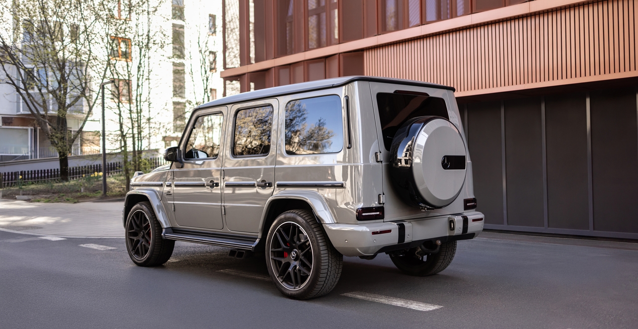 Tył Mercedes-AMG G63 z charakterystycznym pokrowcem na koło zapasowe i potężnymi felgami, na tle miejskiej architektury — styl rozpoznawalny od razu.