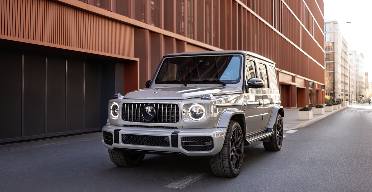 Mercedes G63 AMG na tle nowoczesnej architektury miejskiej, widok z przodu z włączonymi światłami do jazdy dziennej.